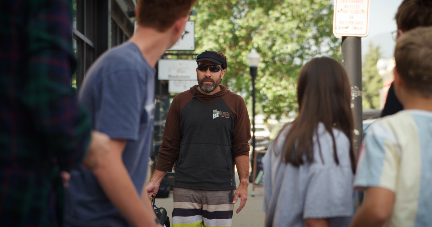 A bearded local in sunglasses talks with a group of teens on Main Street.