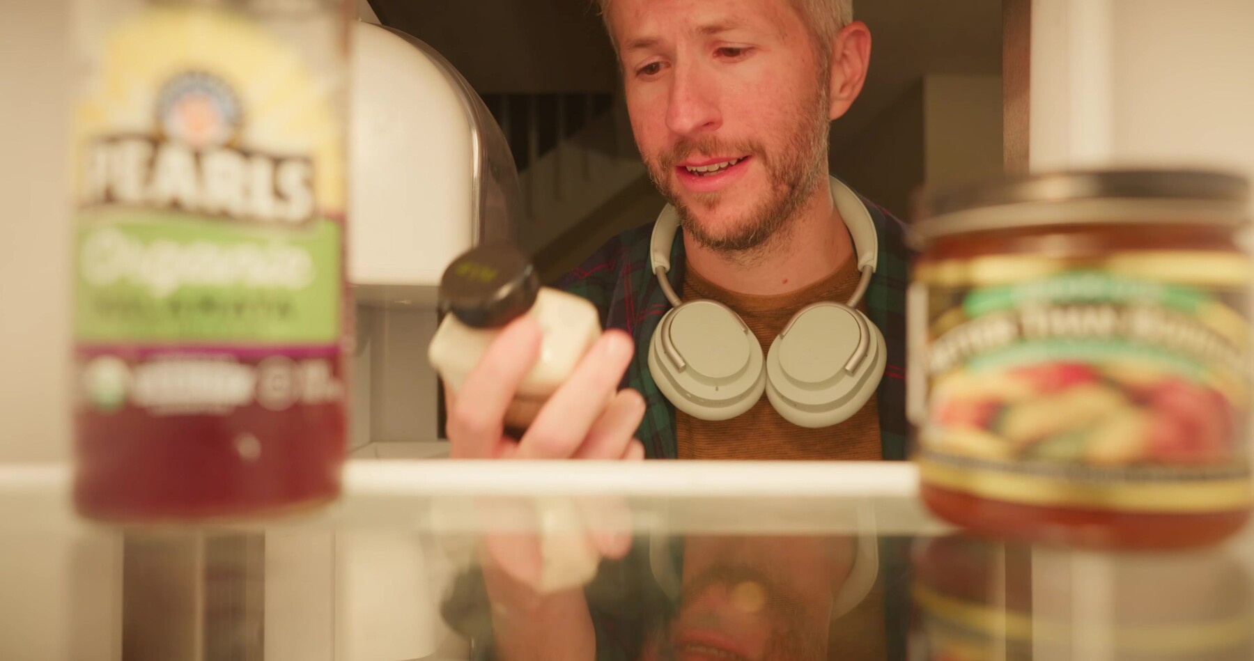 He holds up a small jar from the fridge, examining it, headphones around his neck.