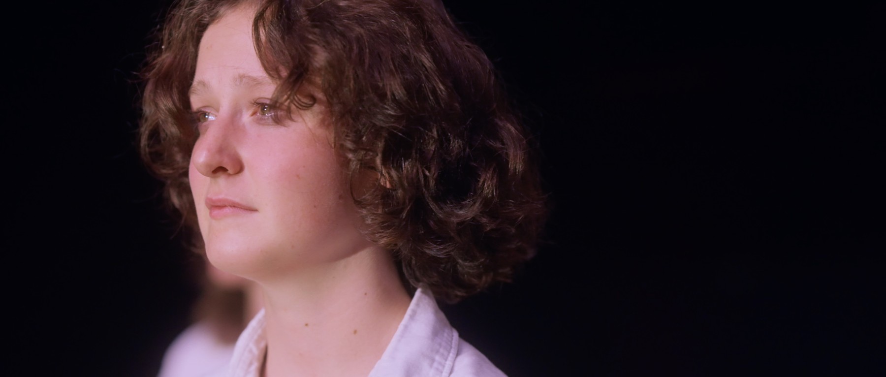Sadie in a white shirt, lit against a dark background.