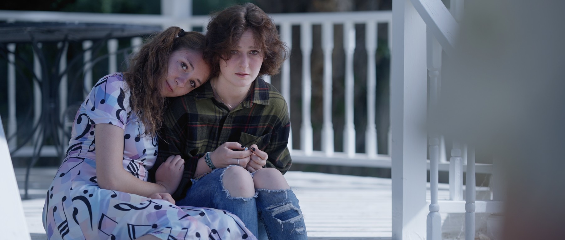 Two young women sit close together on a porch.