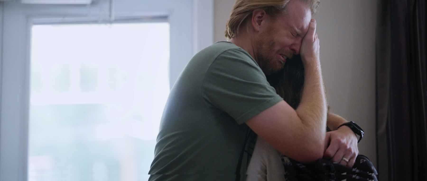 A man embraces a young woman beside a bright window.